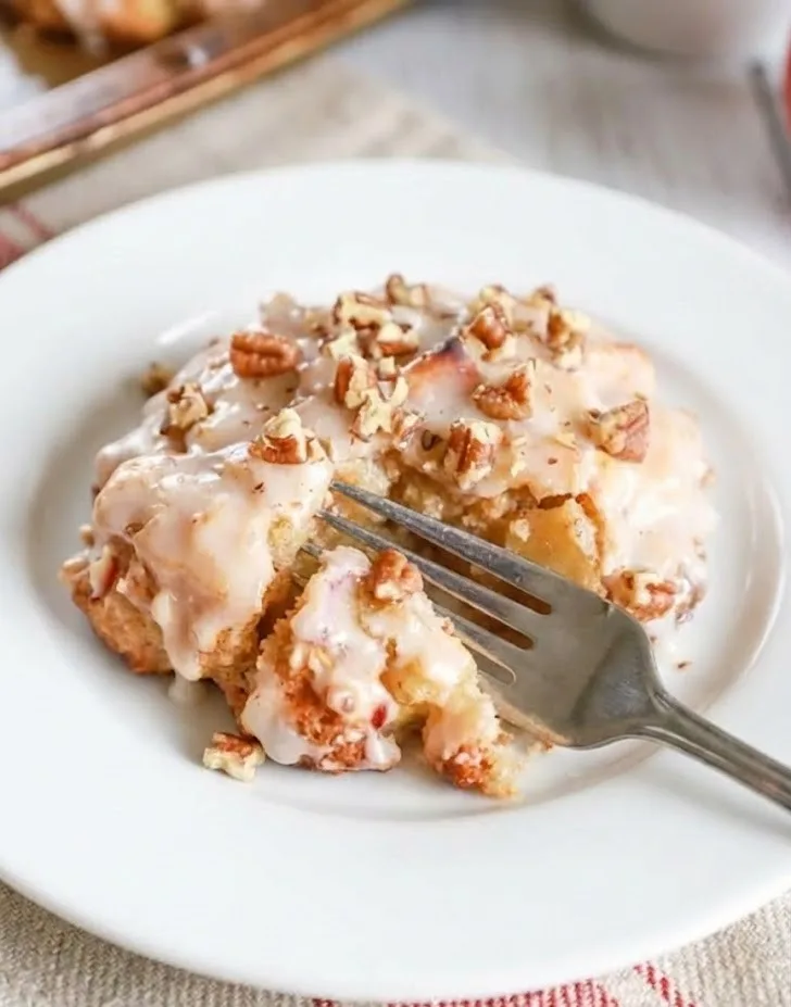 Baked apple fritter with glaze and chopped nuts, partially cut with a fork to show soft apple-filled interior on a white plate
