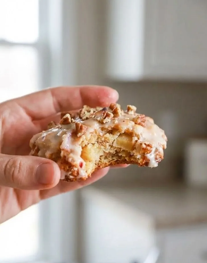 Hand holding a baked apple fritter with a bite taken out, showing soft apple chunks, cinnamon crumb texture, and glazed topping