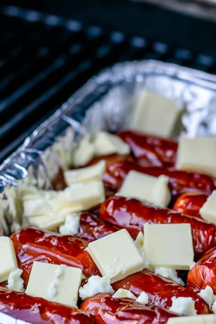 Hot dogs in a foil pan with butter cooking on a grill for hot dog burnt ends