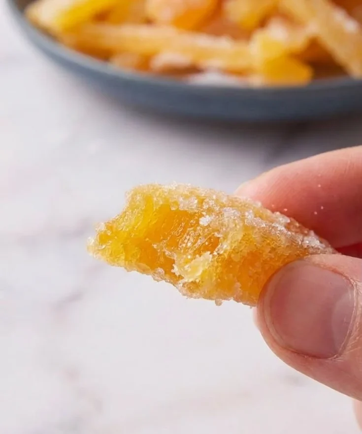 Close-up of a bitten candied orange peel strip coated in sugar held between fingers with blurred bowl in background