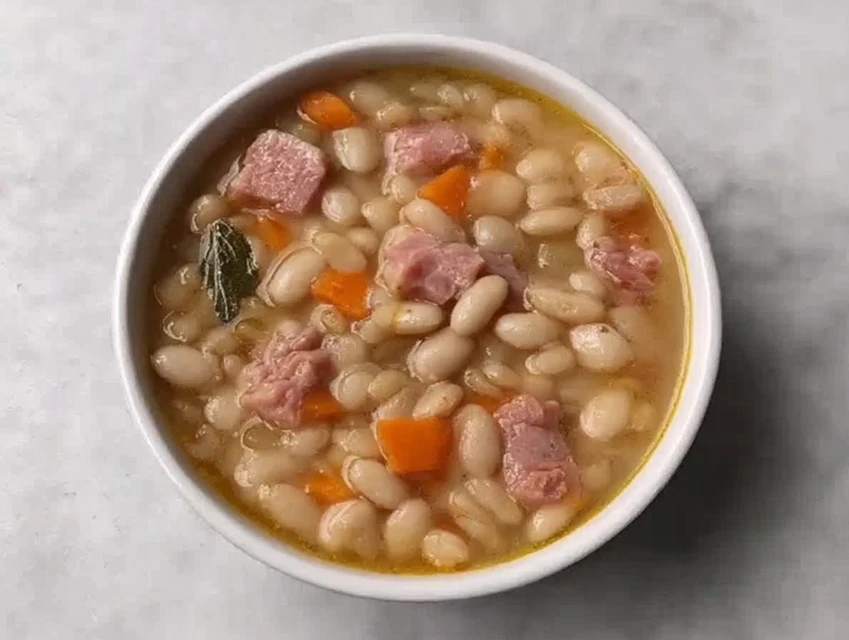 Close-up of homemade Ham And Bean Soup with navy beans, diced ham, carrots, and bay leaf in a white bowl.
