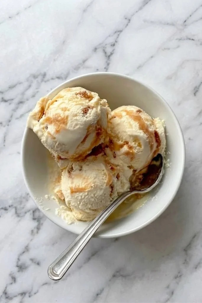 Overhead view of three scoops of honeycomb ice cream in a white bowl showing caramel ripples and crunch.
