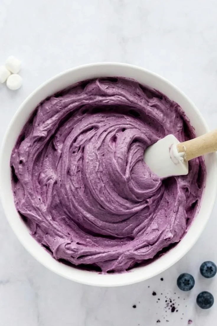 A top-down view of a white bowl filled with thick, swirled purple blueberry marshmallow frosting and a white spatula on a marble surface with fresh blueberries.