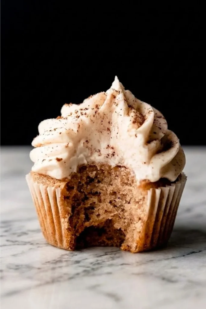 A close-up shot of a spiced cupcake topped with a tall swirl of Chai Spice Buttercream with a bite taken out to show the moist crumb.