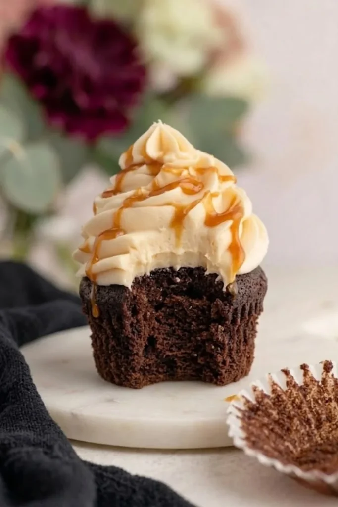 A bitten dark chocolate cupcake topped with a swirl of salted caramel frosting and a generous drizzle of golden caramel sauce.