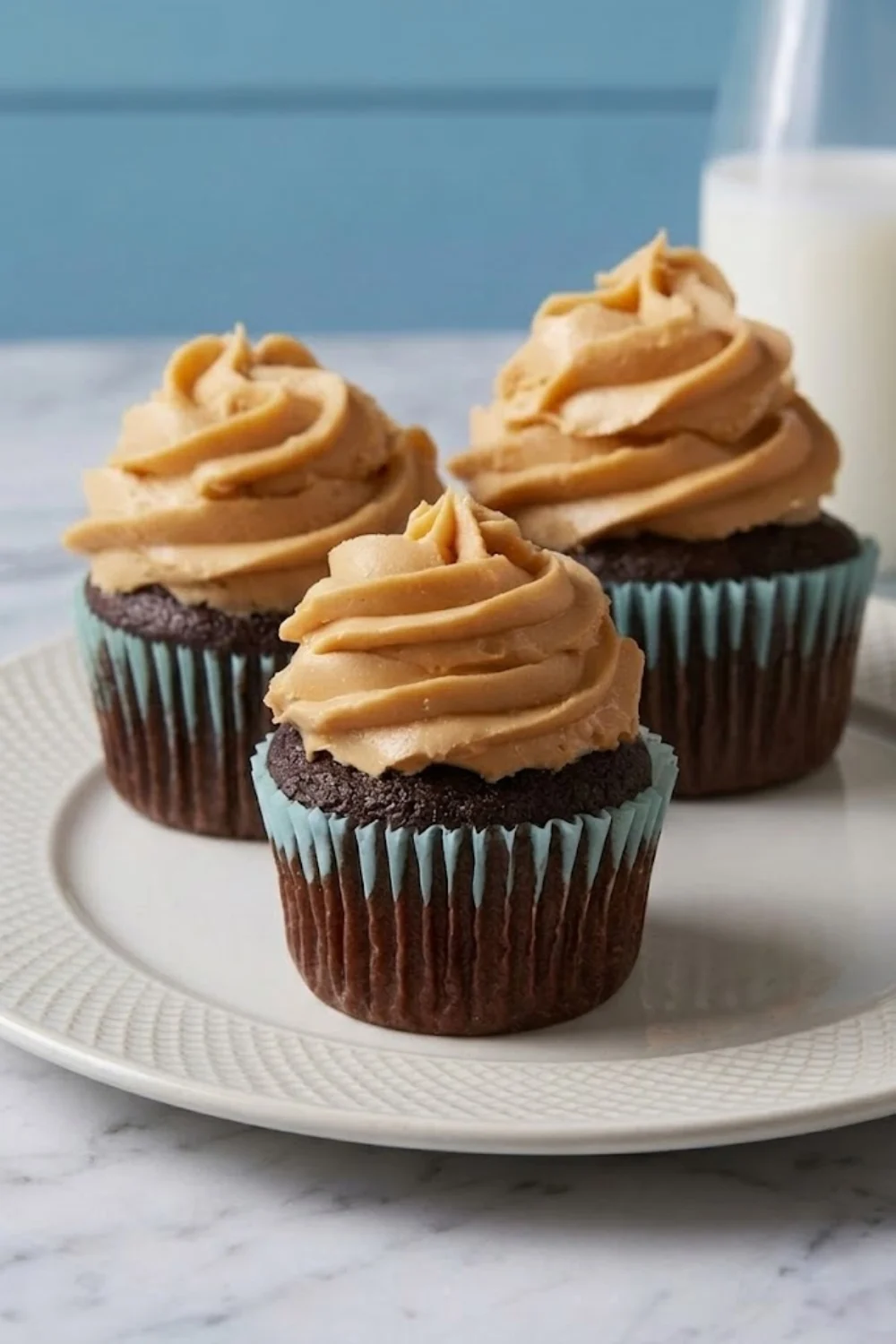 Three dark chocolate cupcakes in light blue and white striped paper liners are arranged on a white ceramic plate. Each cupcake is topped with a tall, perfect, pipeable swirl of creamy peanut butter frosting. A blurry bottle of milk is in the background on a marble surface.