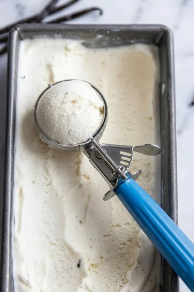 Metal scoop taking a serving of vanilla ice cream from a rectangular metal loaf pan.