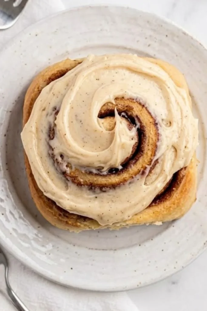 A warm, golden-brown cinnamon roll topped with a generous swirl of melted cream cheese frosting on a white plate.