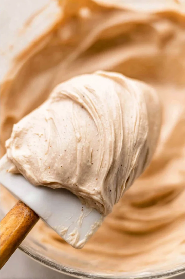 A white rubber spatula holding a large, fluffy dollop of Chai Spice Buttercream over a mixing bowl.