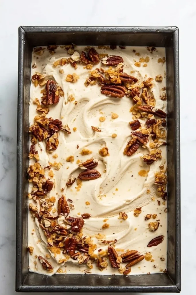 A full metal loaf pan of oatmeal ice cream generously topped with extra toasted walnuts and a cinnamon swirl on a marble background.