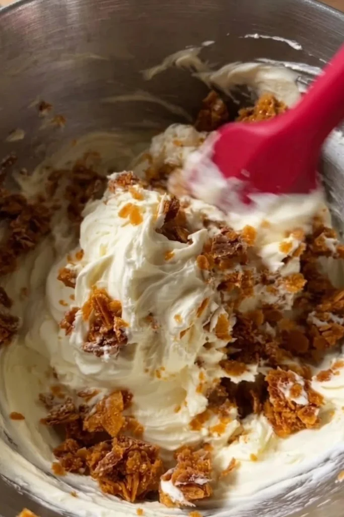 Folding golden honeycomb chunks into a thick, velvety vanilla cream mixture using a red spatula.