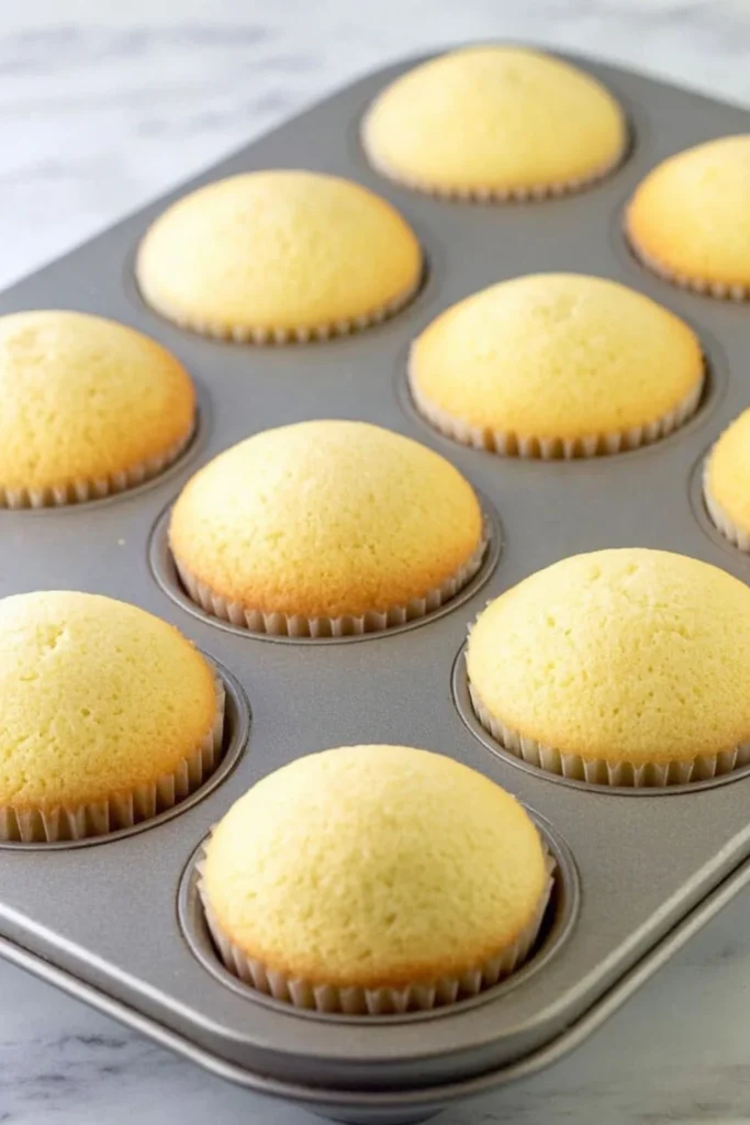 A muffin tin filled with perfectly domed, golden-brown vanilla cup cakes fresh out of the oven.