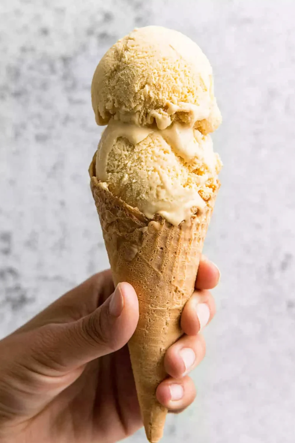 A person holding a waffle cone with two creamy scoops of No-Churn Pumpkin Ice Cream against a neutral background.