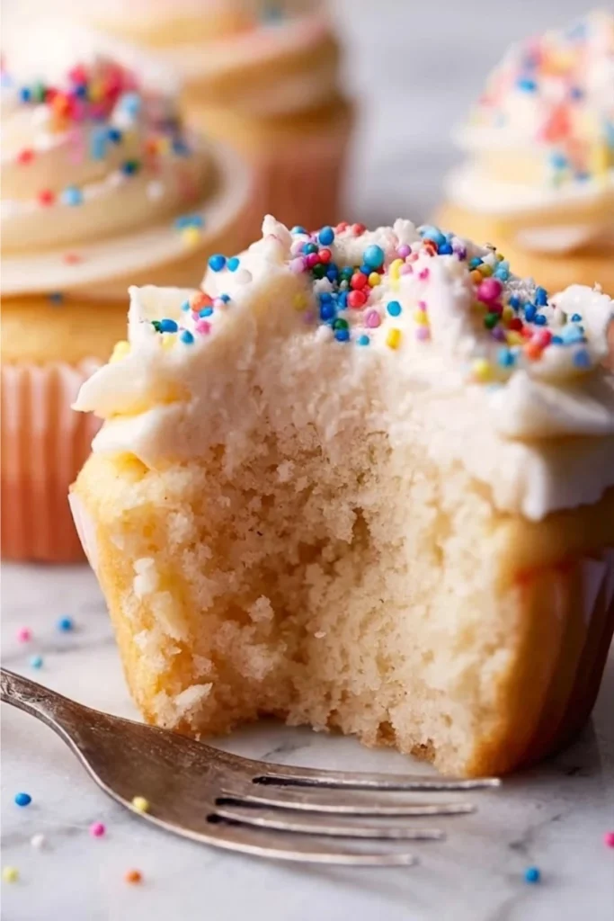 A half-eaten vanilla cupcake showing the soft interior crumb and the creamy layer of fluffy vanilla frosting with sprinkles.