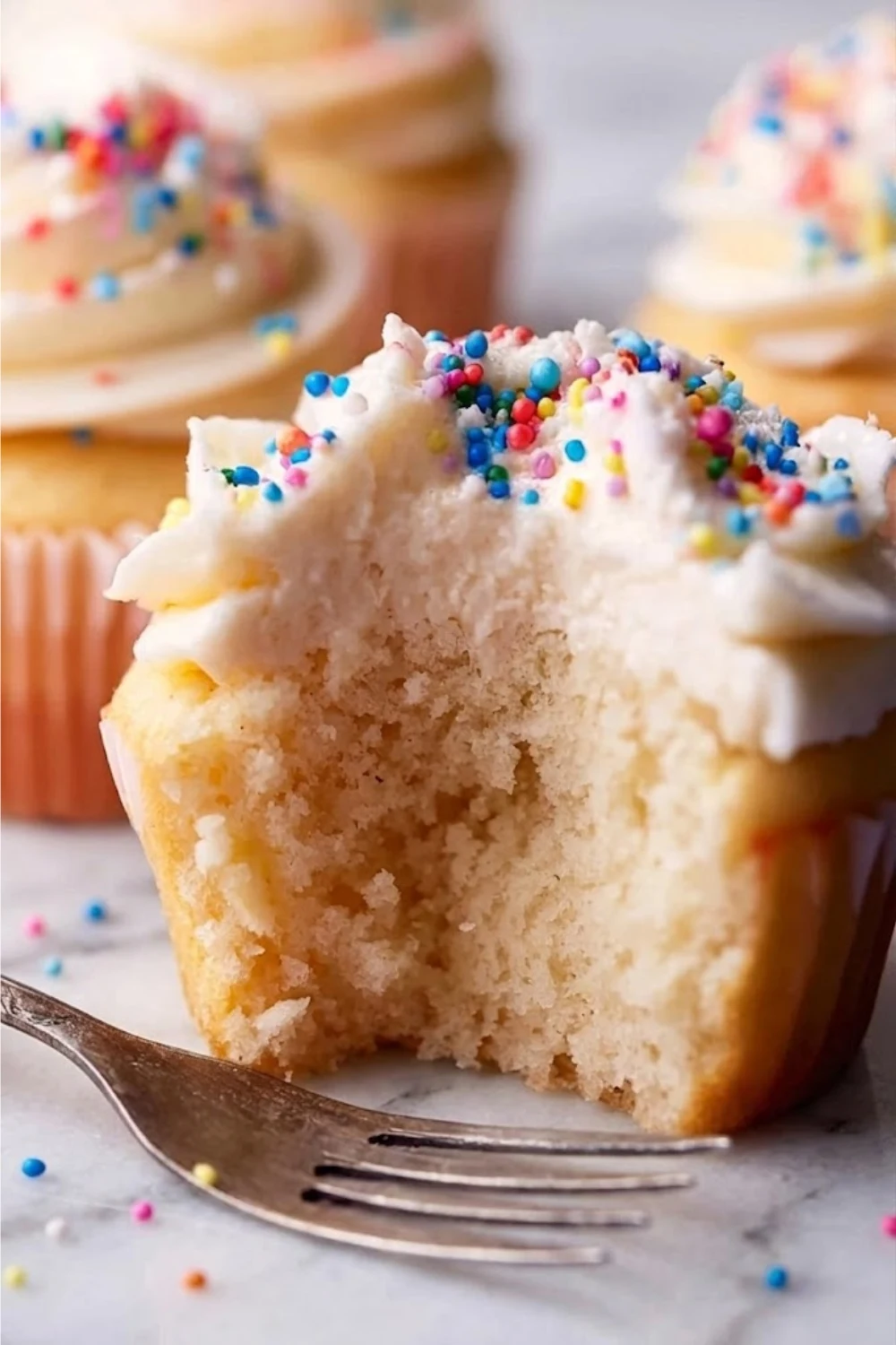 A half-eaten vanilla cupcake showing the soft interior crumb and the creamy layer of fluffy vanilla frosting with sprinkles.