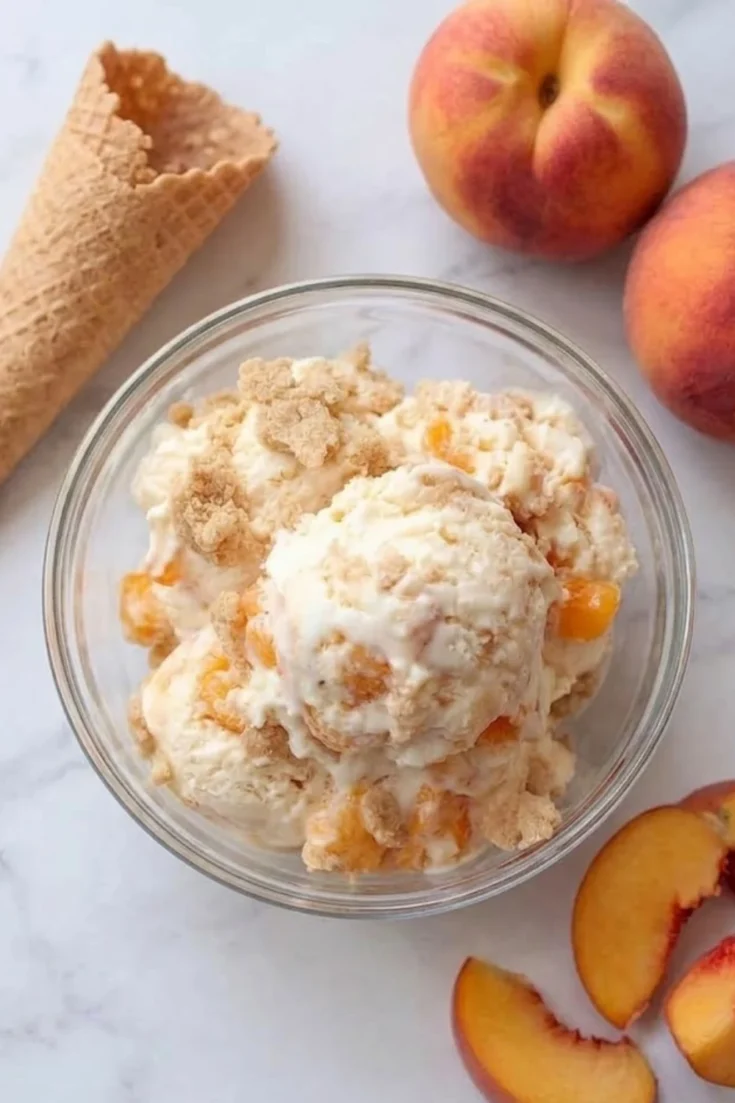 A glass bowl filled with scoops of creamy Peach Pie Ice Cream topped with buttery pie crust crumbles and fresh peach slices.