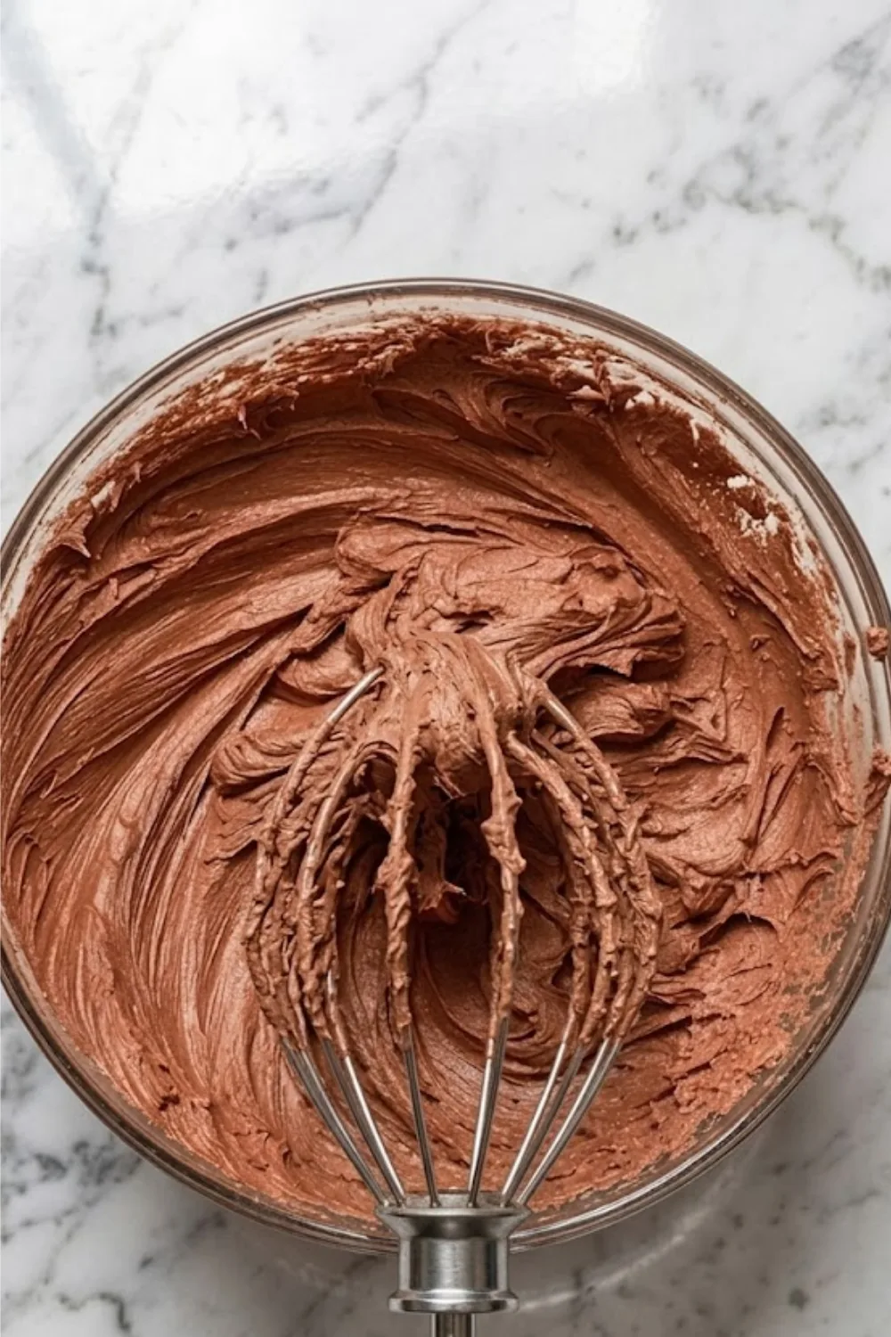 A metal whisk lifting a thick, creamy swirl of chocolate peanut butter frosting from a glass mixing bowl.