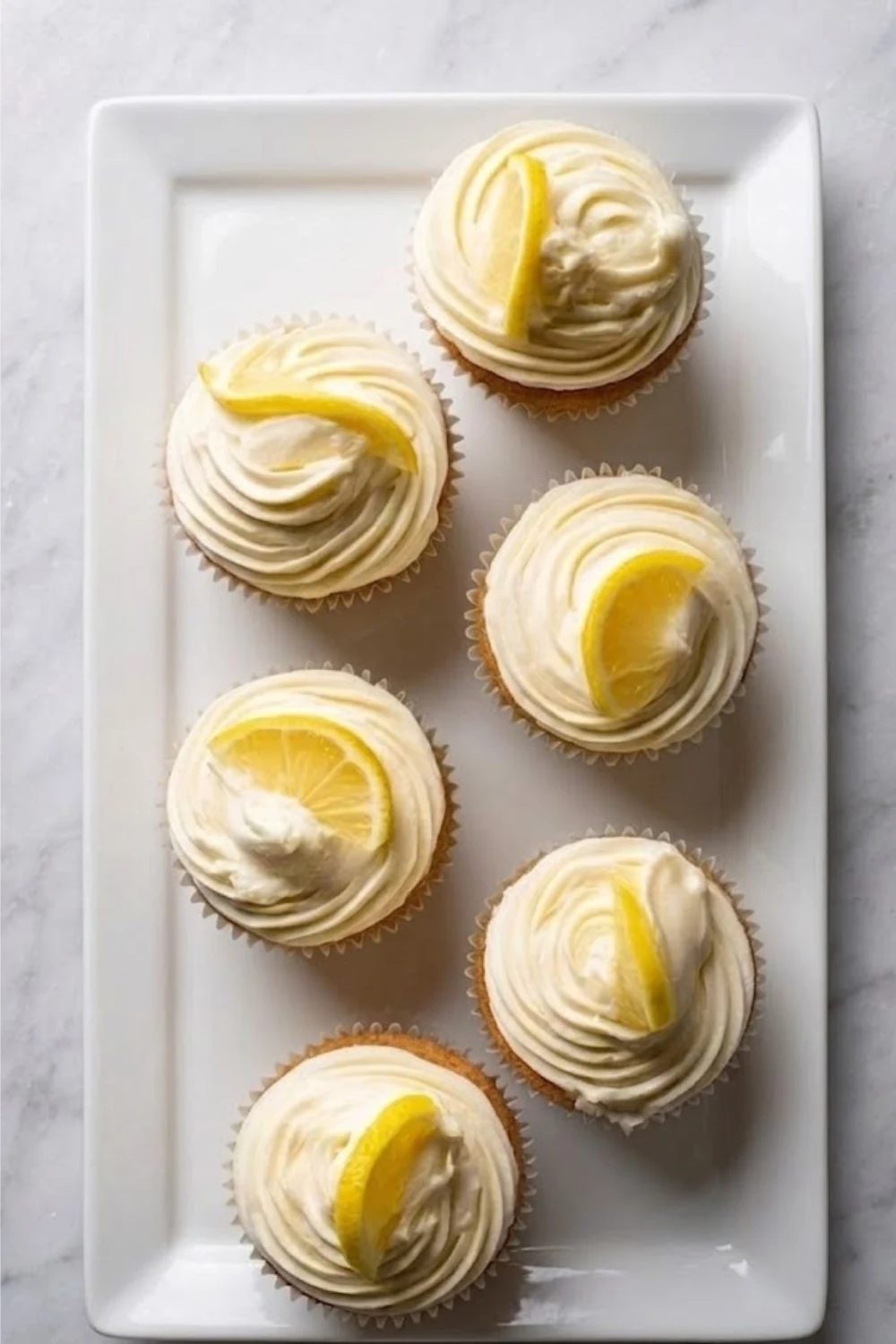 Overhead view of six vanilla cupcakes topped with Lemon Buttercream Frosting swirls and fresh lemon slices on a white rectangular platter.