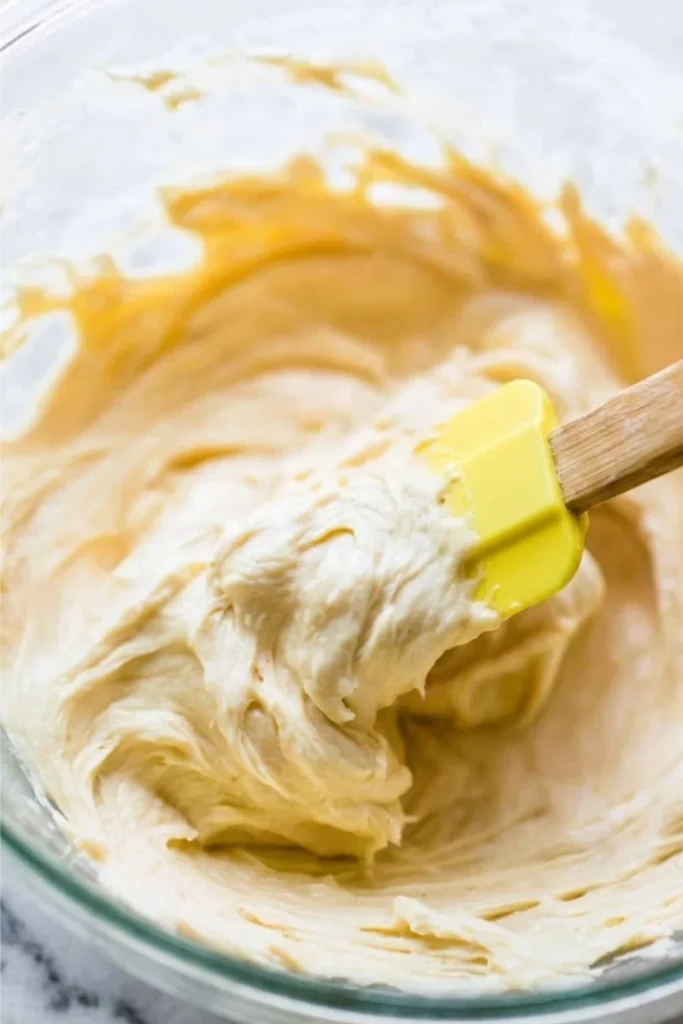 A yellow spatula lifting creamy Lemon Buttercream Frosting out of a glass mixing bowl.