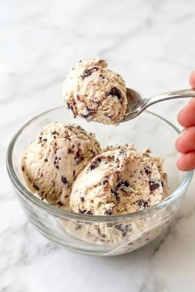 A close-up shot of a hand using a spoon to lift a single, perfectly formed scoop of No-Churn Chocolate Chip Ice Cream from a glass bowl.