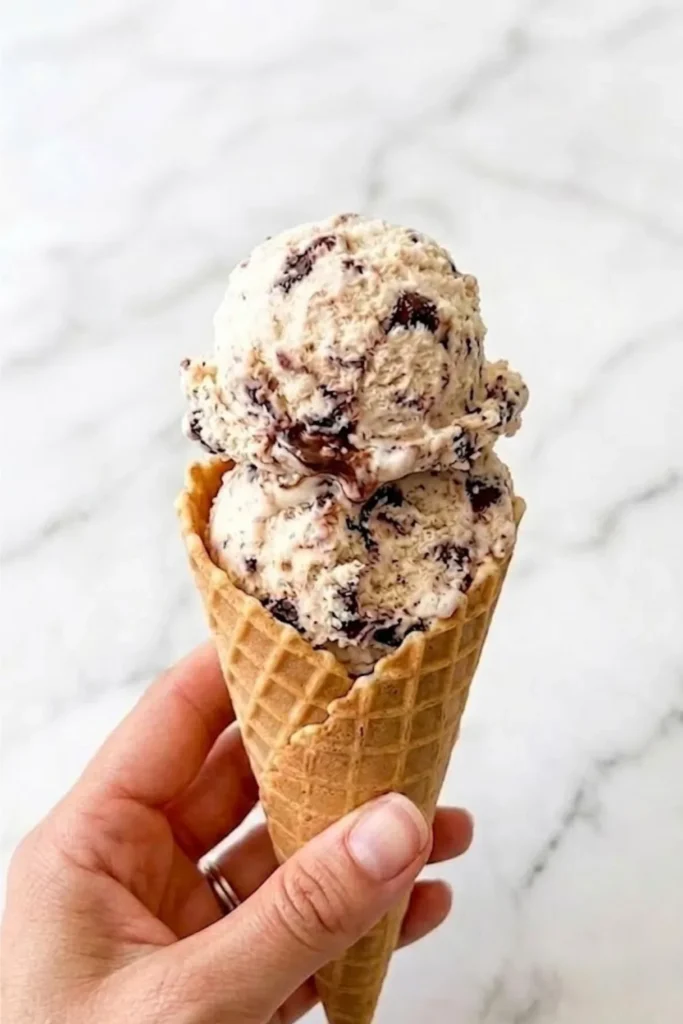 A hand holding a waffle cone piled high with two perfect scoops of No-Churn Chocolate Chip Ice Cream.