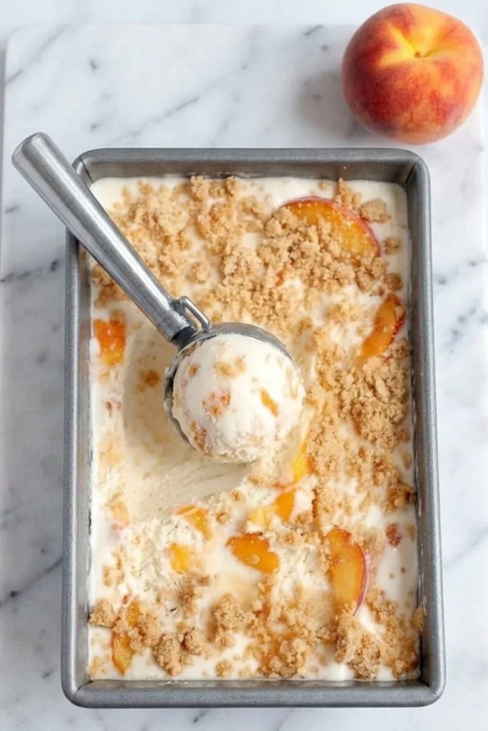An overhead view of a metal loaf pan containing no-churn Peach Pie Ice Cream with a silver scooper lifting a fresh scoop.