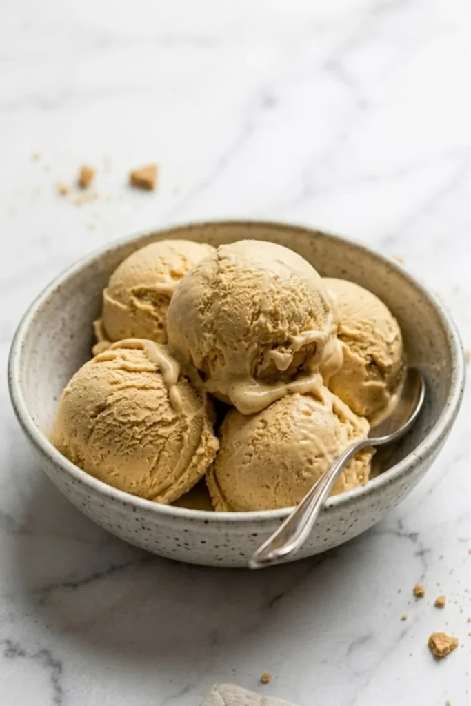 A white speckled bowl filled with several smooth scoops of No-Churn Pumpkin Ice Cream and a silver spoon.