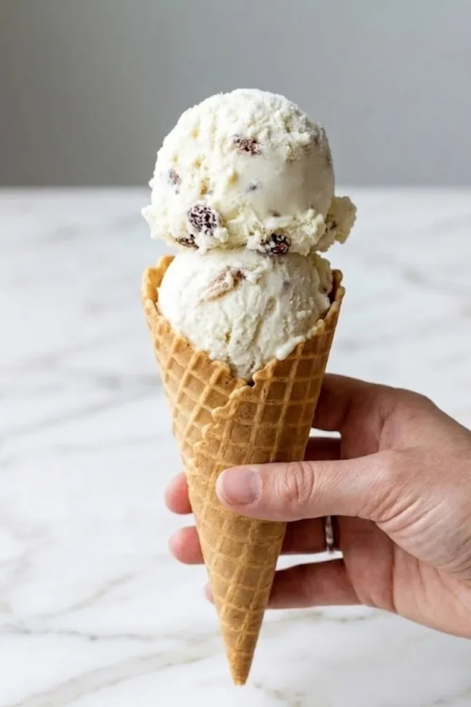 A hand holding a crispy waffle cone with two large scoops of creamy oatmeal ice cream with toasted walnuts.