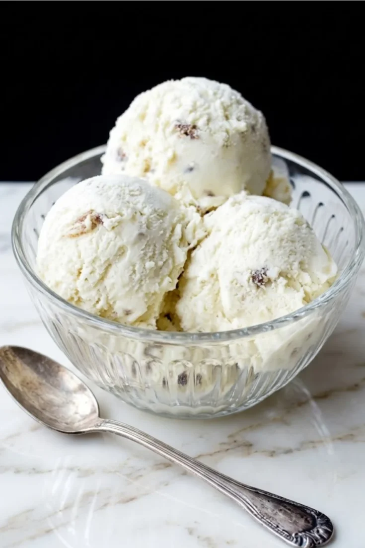 Three scoops of oatmeal ice cream with toasted walnuts served in a clear glass bowl on a marble surface with a vintage silver spoon.