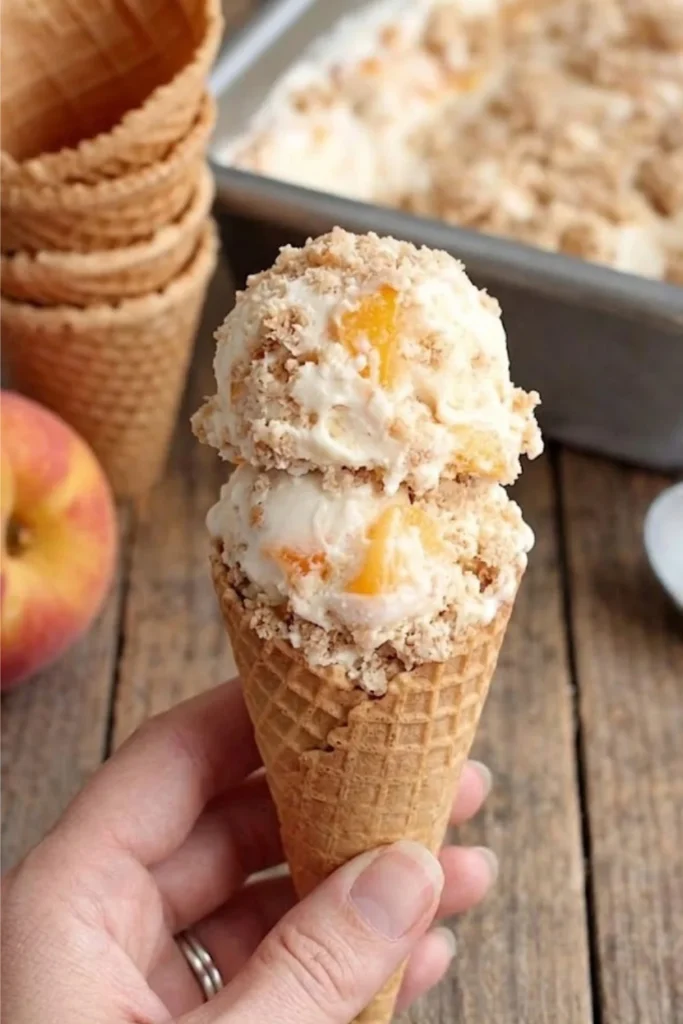 A hand holding a crispy waffle cone with two large scoops of Peach Pie Ice Cream dripping with fresh peach flavor.