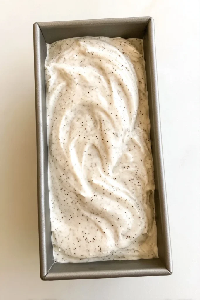 Overhead view of the creamy vanilla base for No-Churn Chocolate Chip Ice Cream with tiny chocolate flecks, sitting unsmoothed in a metal loaf pan before freezing.