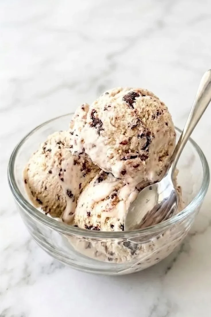 A glass bowl filled with creamy No-Churn Chocolate Chip Ice Cream on a marble background, with a silver spoon resting on the edge of the bowl.