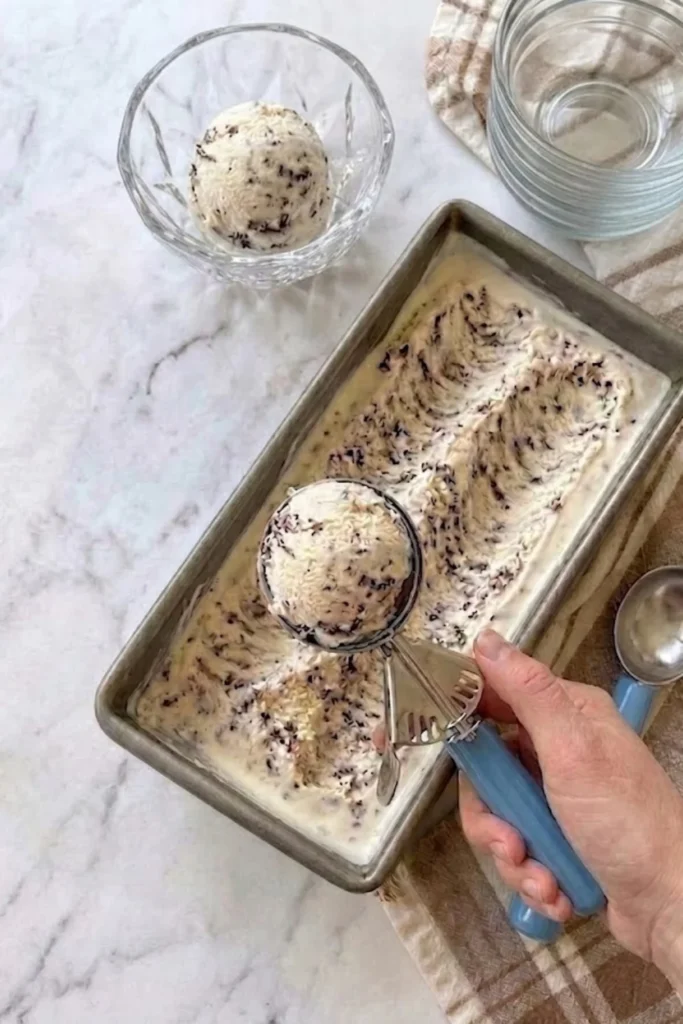 An overhead shot on a marble surface showing a hand using a blue-handled ice cream scoop to remove a scoop of No-Churn Chocolate Chip Ice Cream from a metal loaf pan.