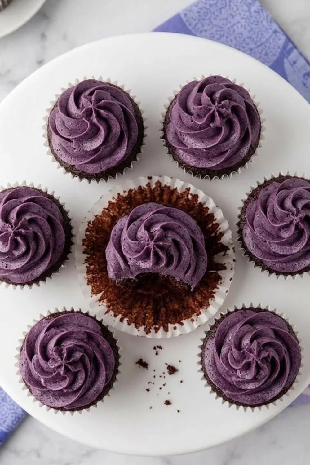 A top-down view of six chocolate cupcakes arranged on a white stand, topped with thick swirls of purple blueberry marshmallow frosting, featuring one cupcake with a bite taken out.